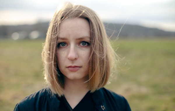 На фото Вера, у нее светлые прямые волосы до плеч, серо-голубые глаза. На щеках золотые блестки и пирсинг в септуме. Она смотрит в кадр, одета в черное.